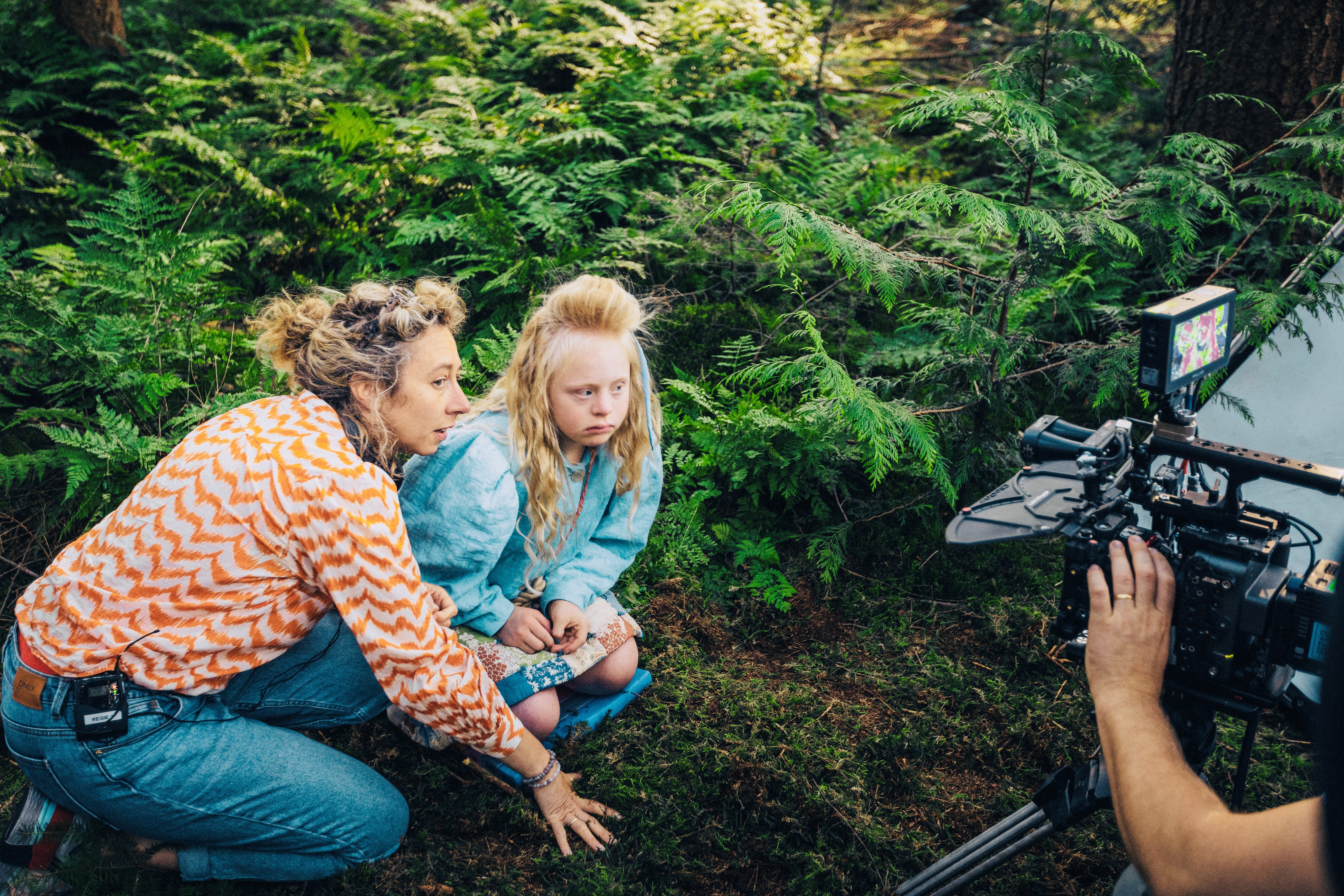 Een vrouw en een kind zitten op de grond in het bos, vanaf rechts staat er een camera op ze gericht