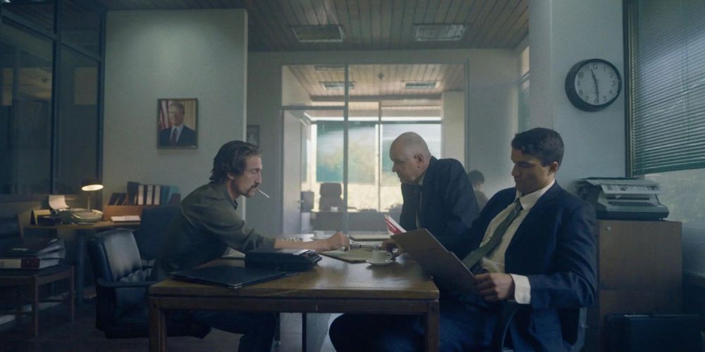 Three men sit at a table, one of them is reading a file.